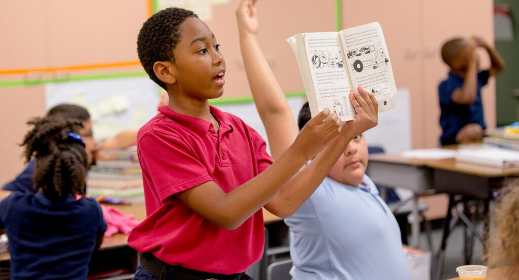 A young boy with short, curly hair and dark skin holds up a book in a classroom.