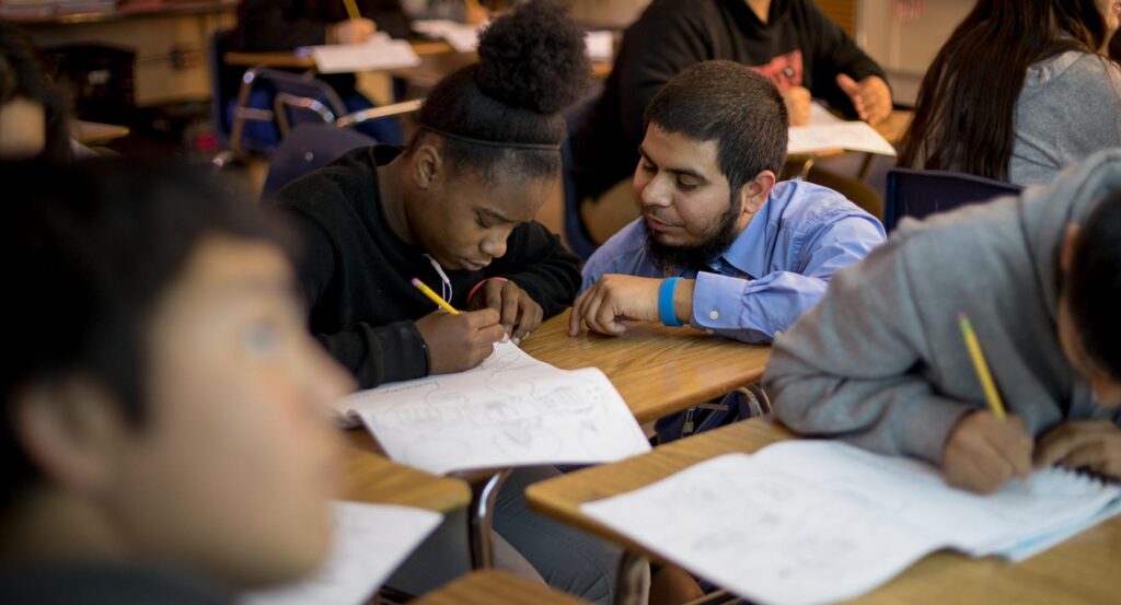 A Latino teacher works with a black teenaged girl on an assignment in a classroom full of diversity.