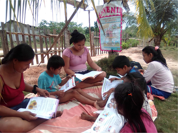 Photo of a group of young children sitting outside with their teacher reading books.