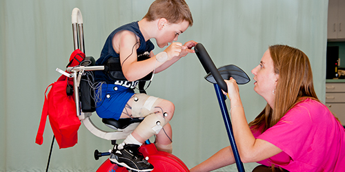 A young woman works with a young boy receiving physical therapy on an exercise bike.