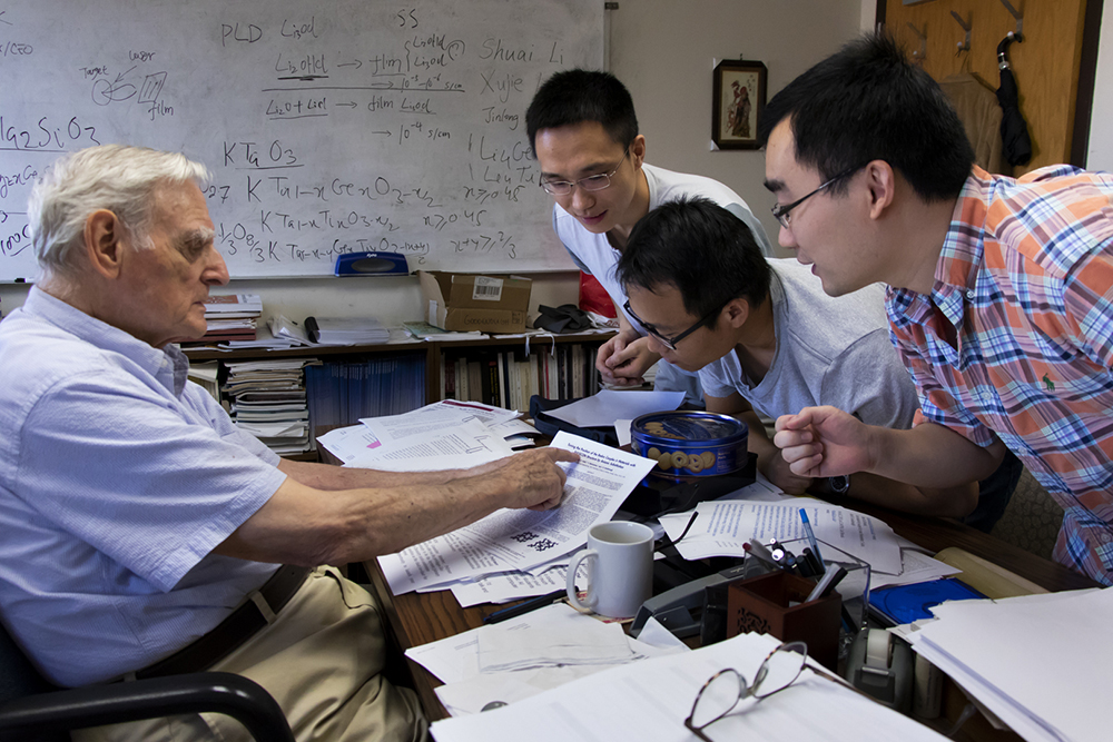 Professor John B. Goodenough sits at his desk while reviewing a paper with three students. 